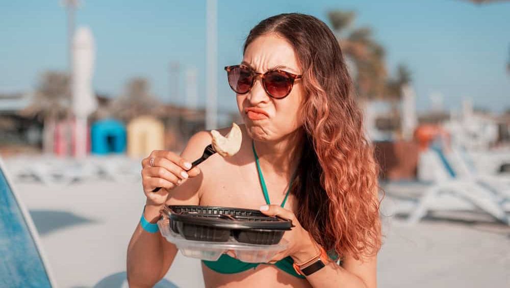 Tourist grimacing while eating takeaway food on a beach, expressing disgust and disappointment with the meal