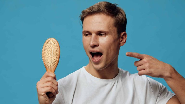 Young man with short brown hair, wearing a plain white t-shirt, holding a wooden hairbrush and pointing at his head with an expressive surprised look isolated on a bright blue background, studio shot
