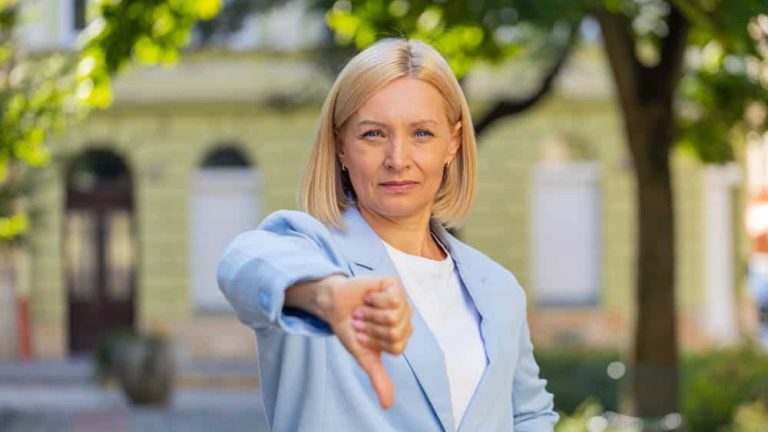 Sad upset disappointed Caucasian mature businesswoman female woman in formal suit showing thumb down sign gesture, rejection finger dislike negative reaction disagreement outdoors town city street.