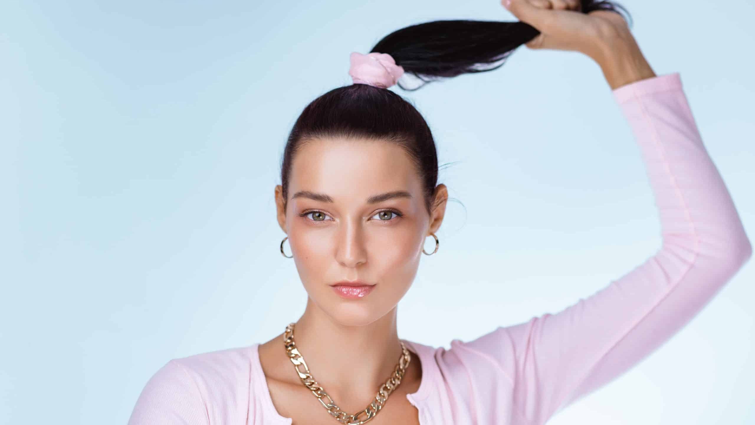 Caucasian woman in modern pink cardigan and gold chain, holding her ponytail. High quality studio fashion concept blending sporty elegance and luxury beauty for branding or ads.