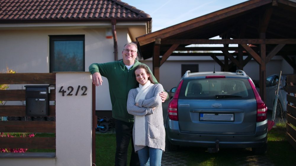 Couple standing together in front of house, man leaning casually on fence while embracing woman, family bond captured outside home with parked car visible in background