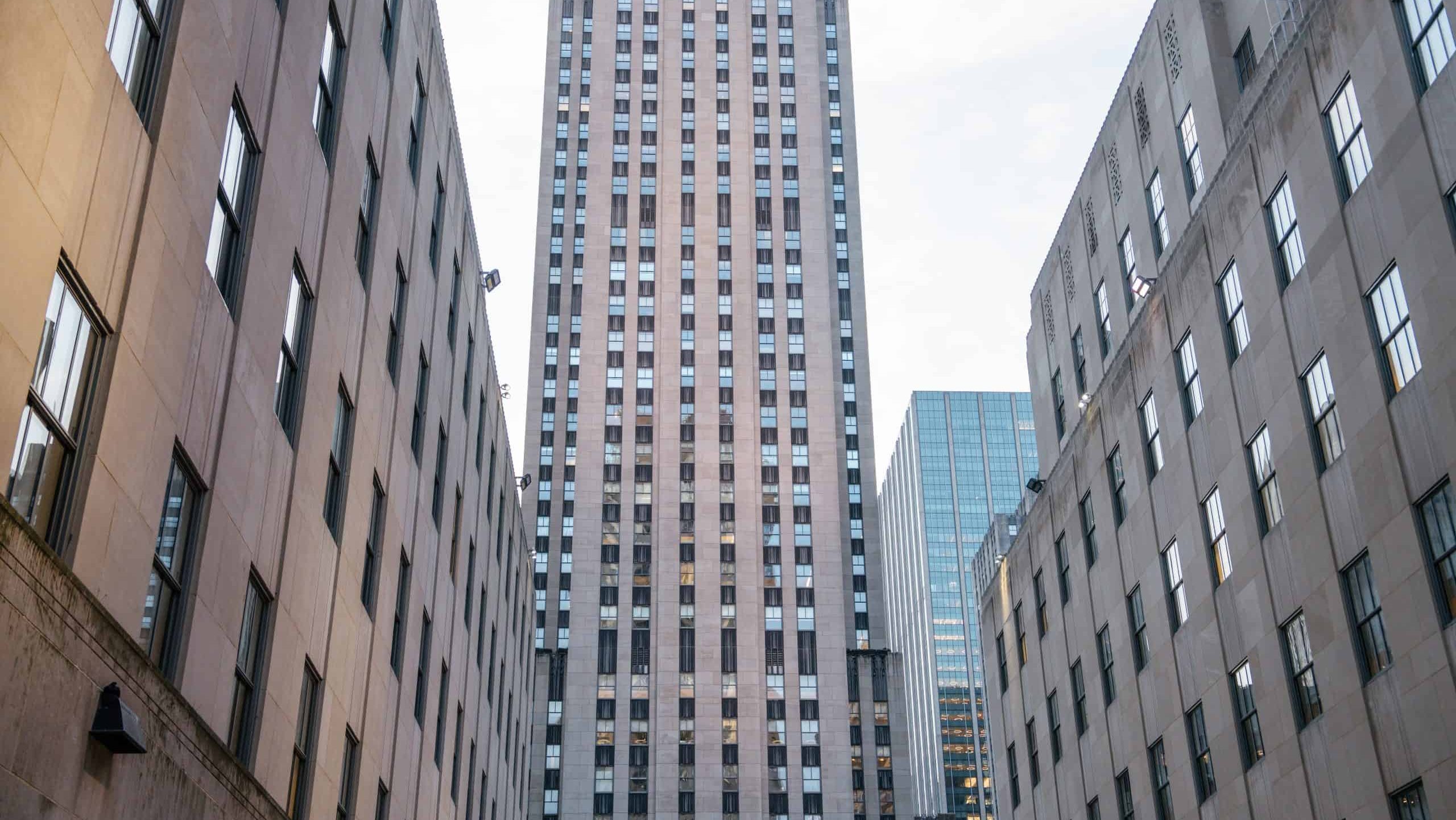 A centered view of the Rockefeller Center tower, framed tightly by symmetrical stone office buildings. The image highlights the Art Deco design and urban density of Midtown Manhattan.