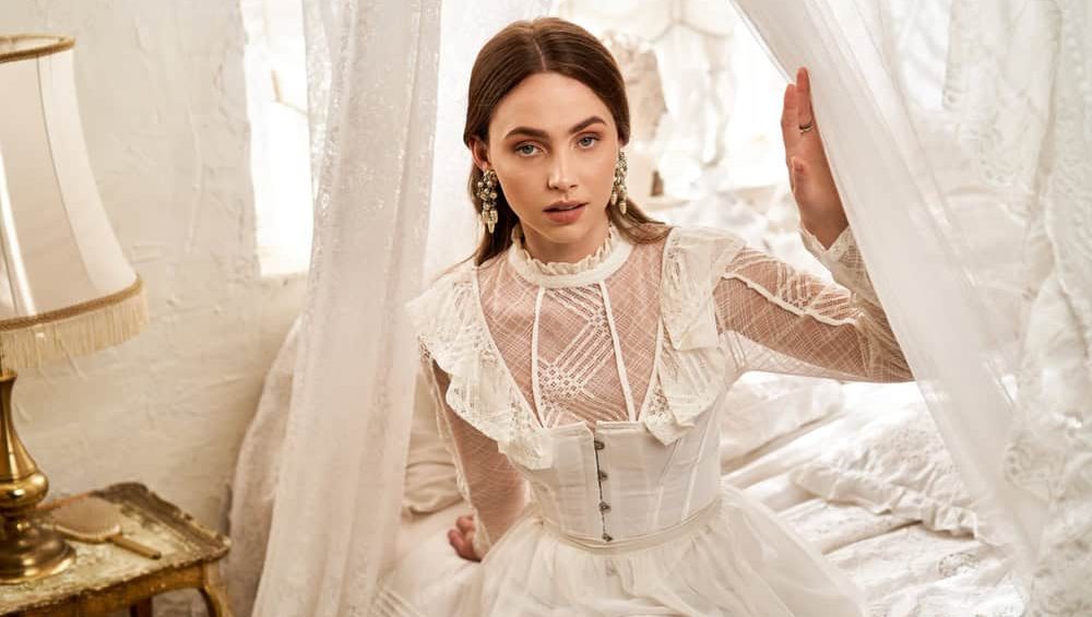 A beautiful young woman in a vintage dress sits elegantly, surrounded by soft, flowing fabrics.