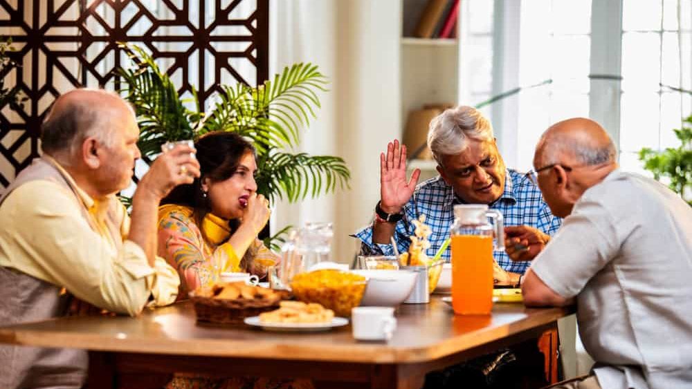 Seniors chatting while having food, Indian Asian old age friends reuniting at home sharing snacks, tea, coffee and meals while engaging in cheerful conversations and laughter around dining table