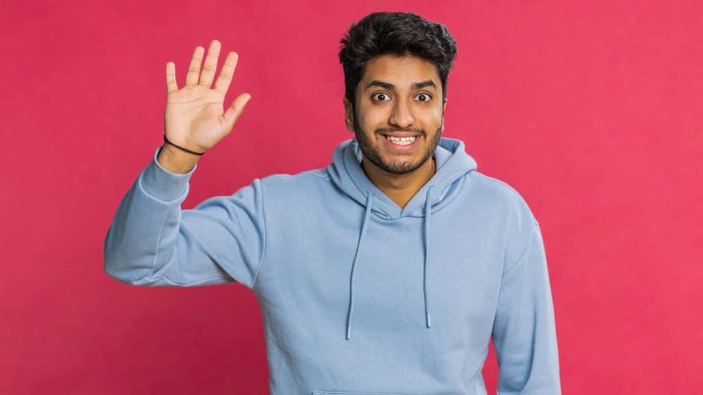 Young Indian man smiling friendly at camera, waving hands gesturing hello greeting or goodbye welcoming with invitation hospitable expression. Arabian Hindu guy isolated on red studio background