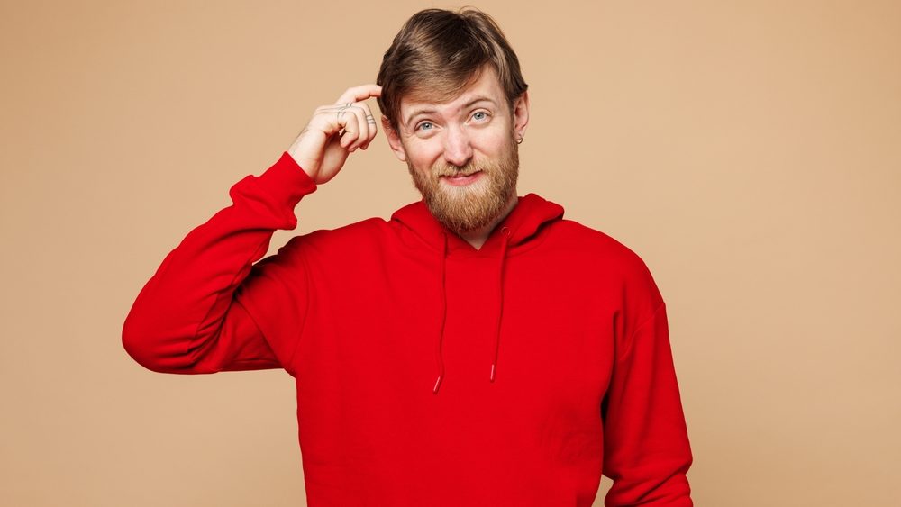 Young mistaken confused sad Caucasian man he wear red hoodie casual clothes look camera scratch head looking camera isolated on pastel plain light beige background studio portrait. Lifestyle concept