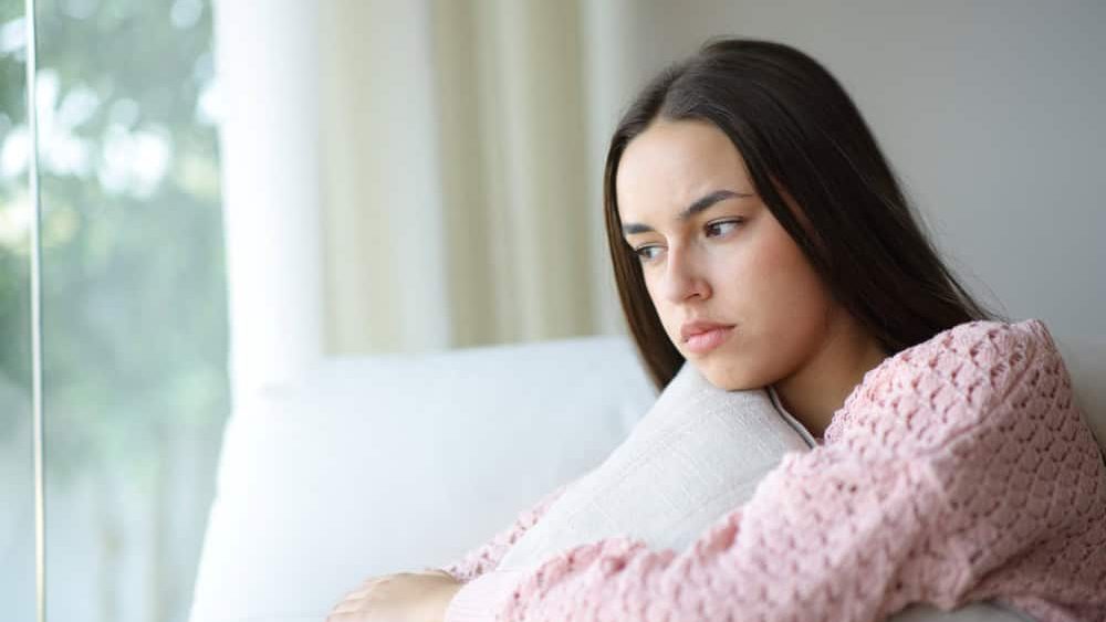 Angry pensive woman looks away through a window sitting on a couch at home