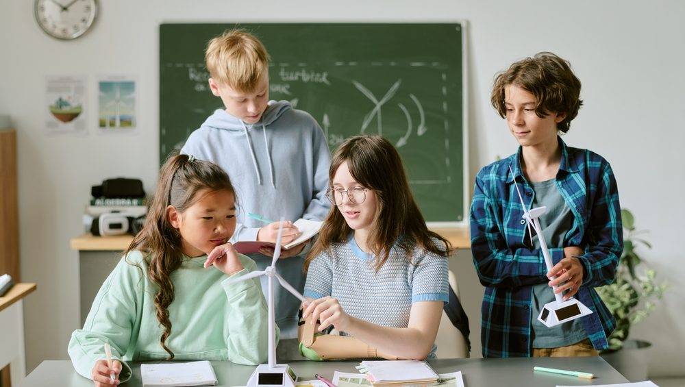 Group of multiethnic teenagers collaborating on science project, assembling wind turbine models and discussing renewable energy concepts in classroom with educational materials visible