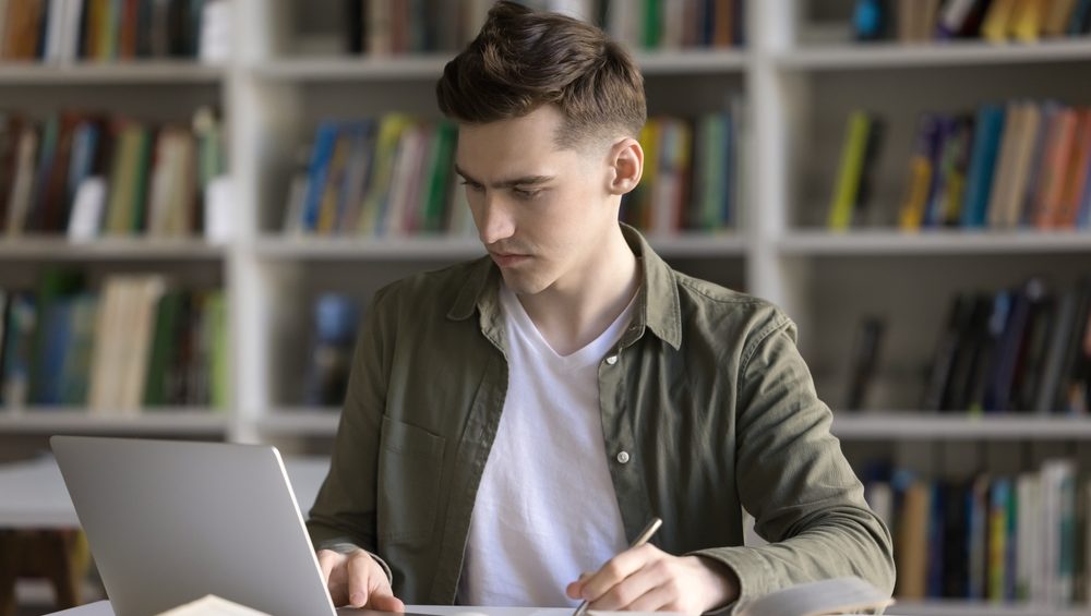 Hybrid learning. Serious focused gen z male student engaged in self training at library desk use notebook paper books to search information write essay assignment thesis diploma project draft by hand