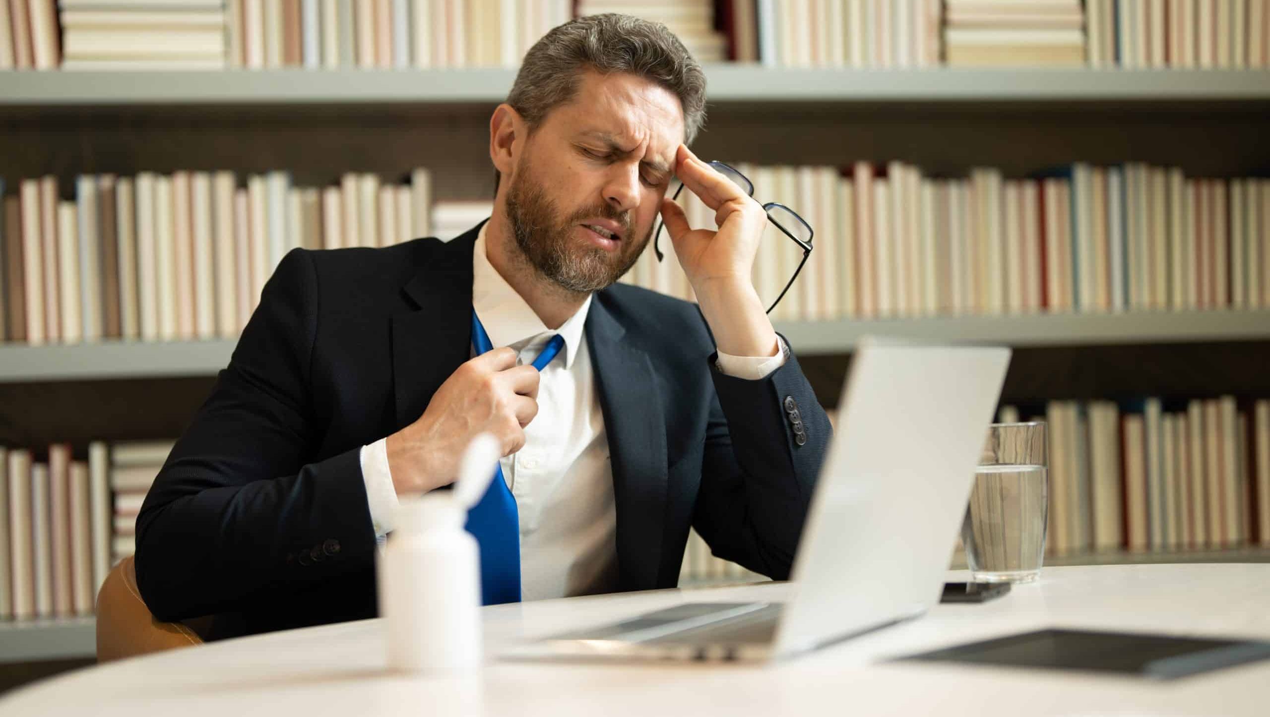 Millennial Businessman in office having headache. Business man in suit work on laptop at home office take off glasses suffer from migraine or headache. Tired exhausted man suffering from headache.