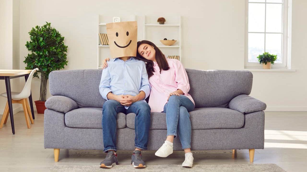 A paper bag with a smile drawn on a man's head, a woman hugging him. A married couple is sitting on the couch at home, hugging each other. Pretense in marriage, reconciliation in relationships