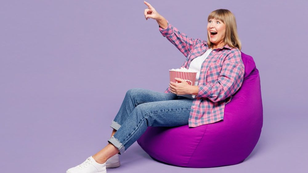 Full body elderly blonde woman 50s years old wear violet shirt casual clothes sit in bag chair watch movie film eat popcorn point finger aside isolated on plain purple background. Lifestyle concept