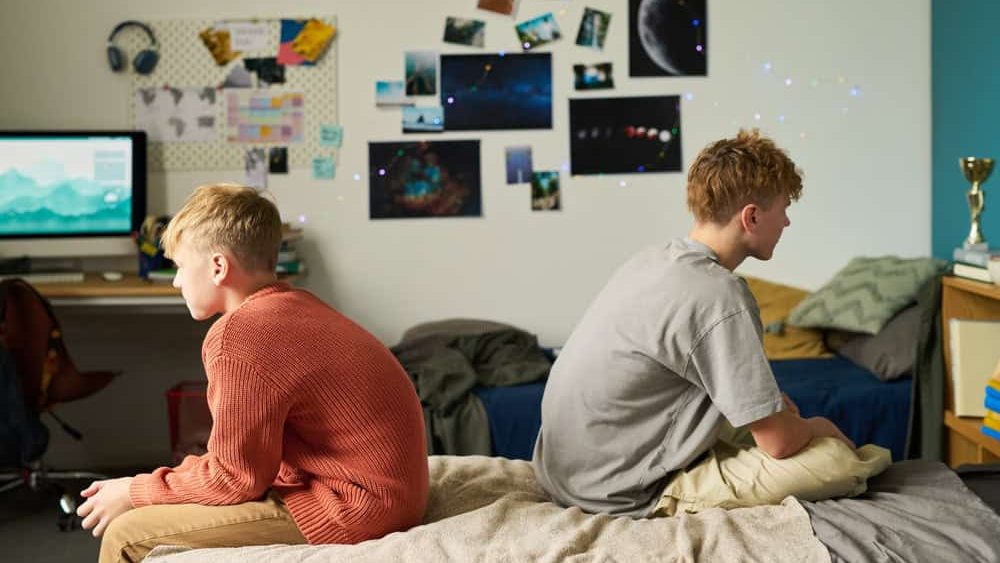 Two Caucasian teenage boys sitting back to back on bed in shared bedroom, both looking away from each other, surrounded by personal items and wall decorations, showing sibling relationship