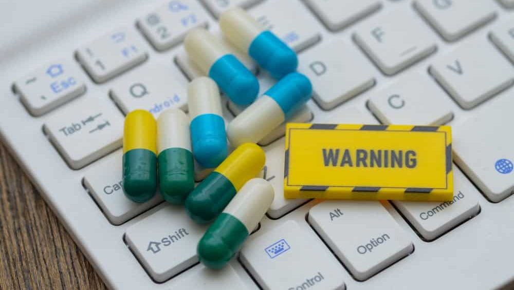A warning sign and scattered pills on a keyboard, symbolizing the risks of buying medication on the internet and counterfeit drugs.