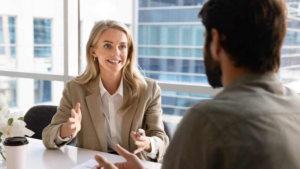 Happy blonde young businesswoman talking to business partner at office table, consulting expert, advisor, asking for expertise, discussing new partnership, teamwork