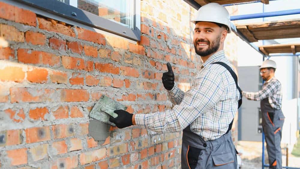 Construction worker wearing hard hat and uniform, smiling at camera, giving thumbs up while plastering brick wall