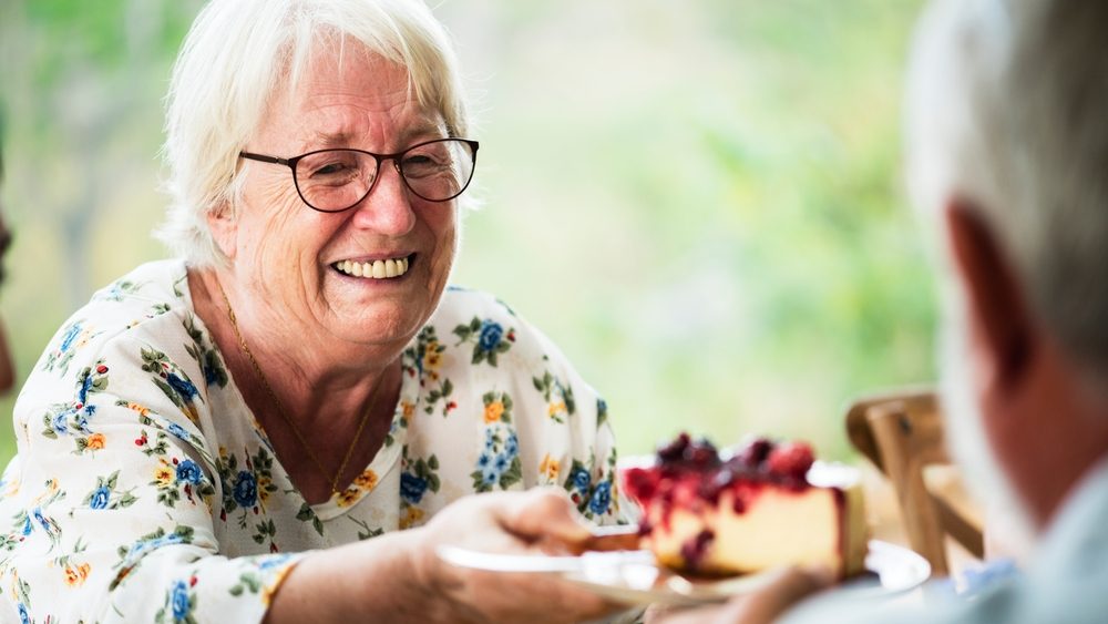 Elderly woman with glasses, smiling, offering a slice of cake. Outdoor setting, cheerful mood. Elderly woman enjoying dessert, sharing cake, happy atmosphere. Christmas food image.