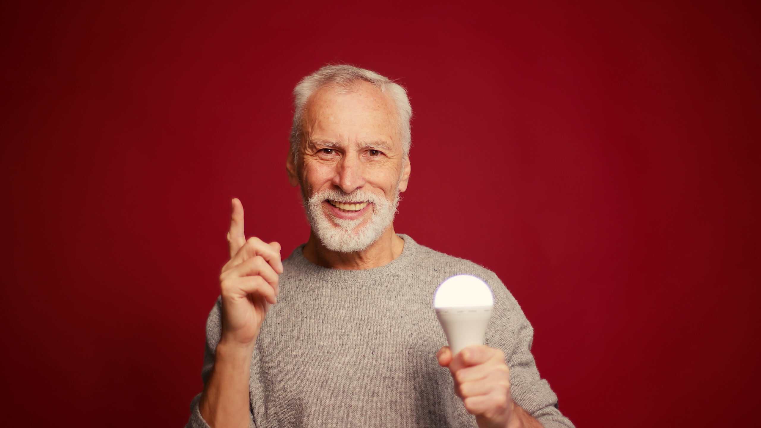 Portrait of senior gray haired man holding light bulb having good idea pointing finger up on red background. Brainstorming concept