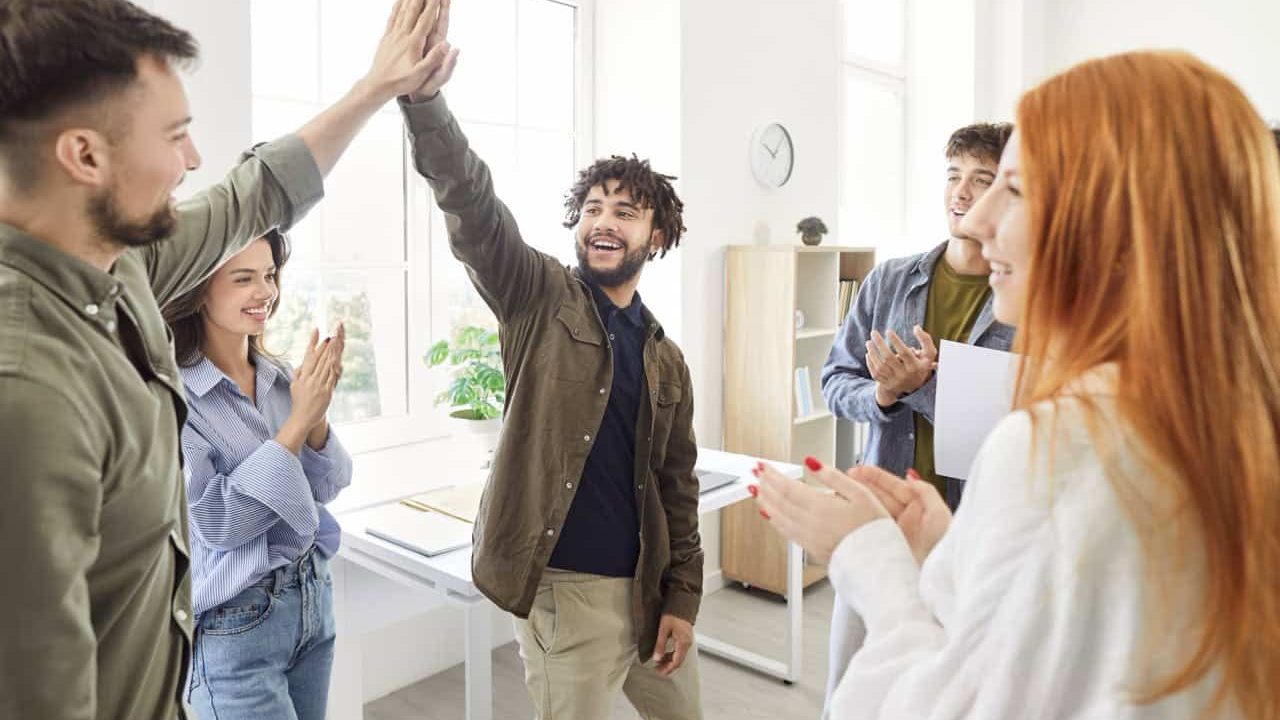 Two men giving high five each other celebrating success, making deal, business achievement or signing contract with group of young people in casual clothes applauding to coworkers in office