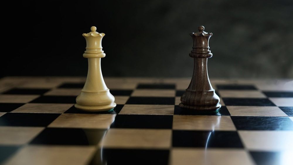 Two opposing chess queen pieces stand facing each other on a black and white chessboard under dramatic lighting.