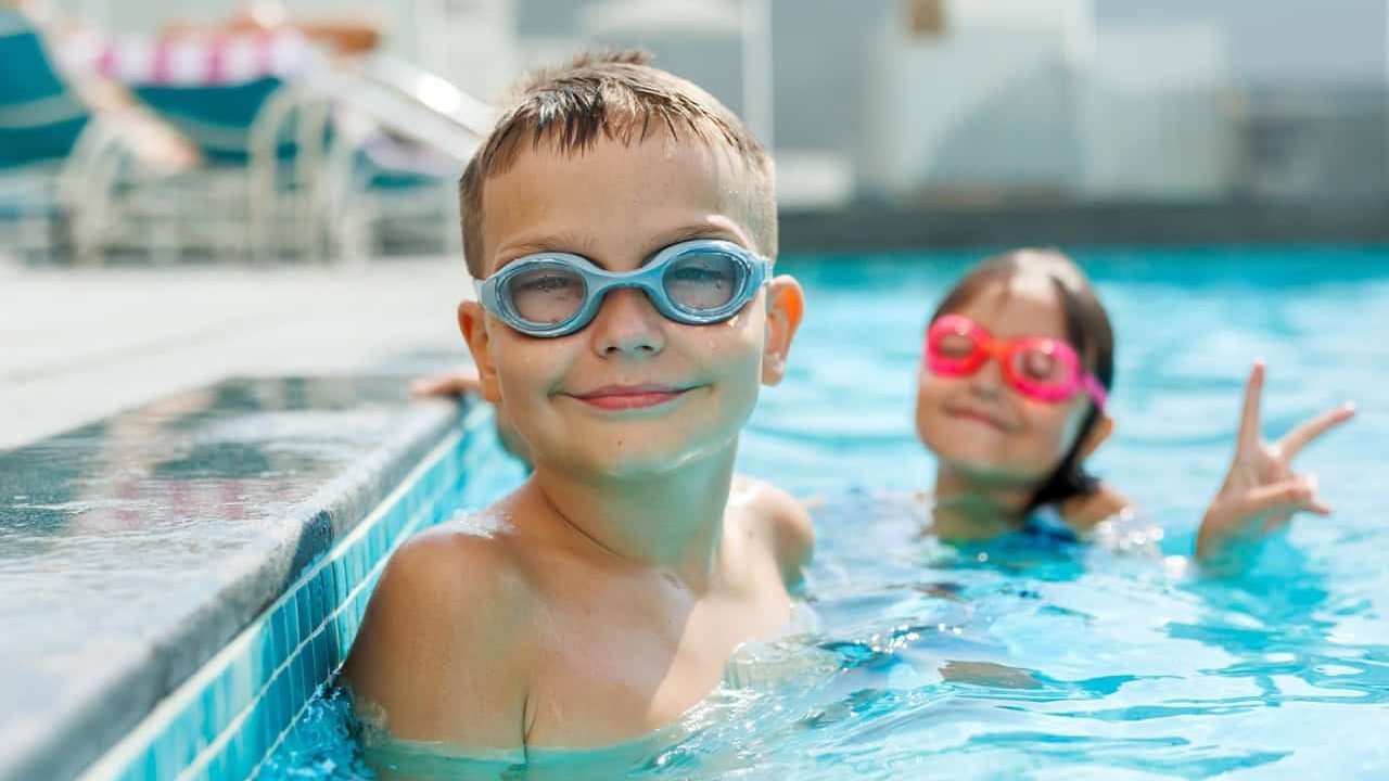 Happy boy swimming in a pool with blue goggles, while little girl smiles in background. Joyful summer vacation for kids.