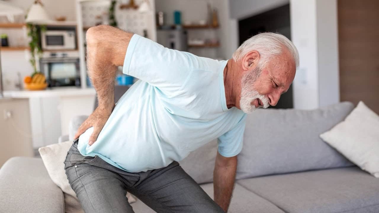 Back pain in the elderly. Senior man holding painful lower back, people with body-muscles problem, healthcare and medicine concept. Retired male standing in the living room and feeling backache.