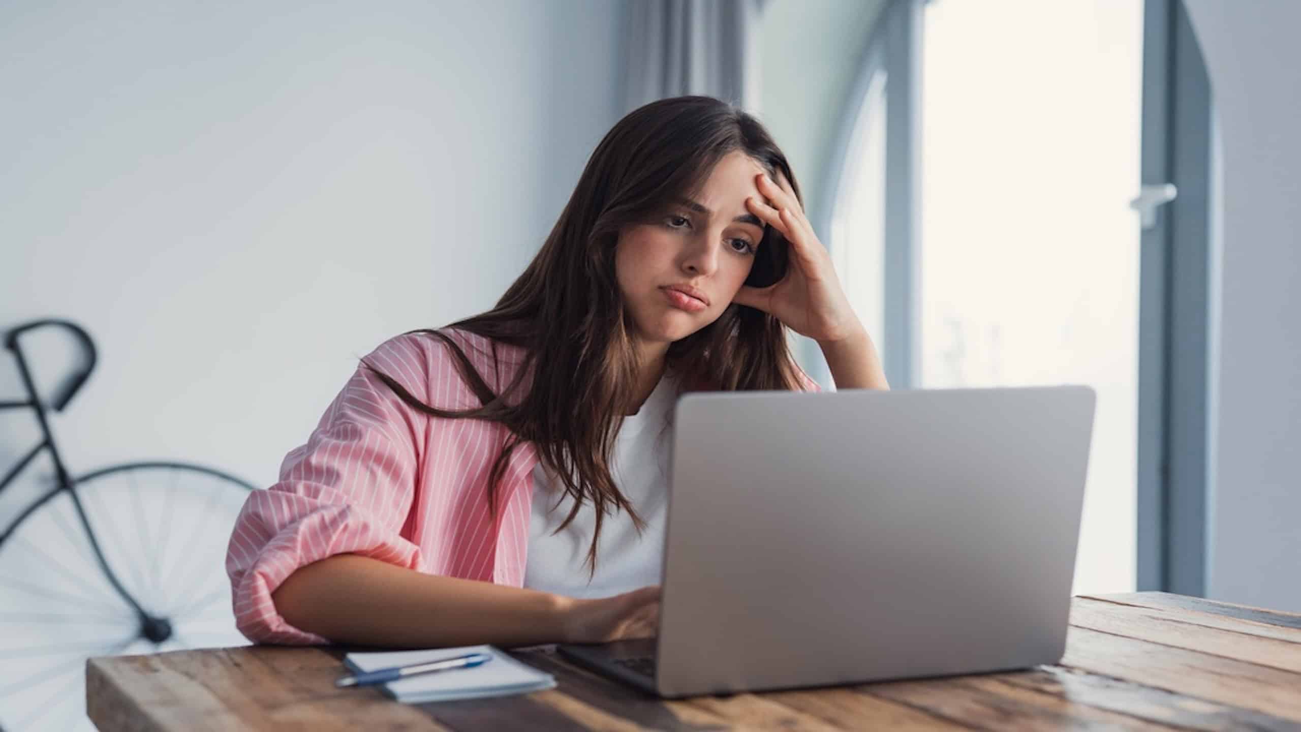 A stressed woman at her desk holds her head in frustration after receiving an unpleasant email, reflecting dismissal fears, financial pressure, and the mental strain of balancing work and personal life