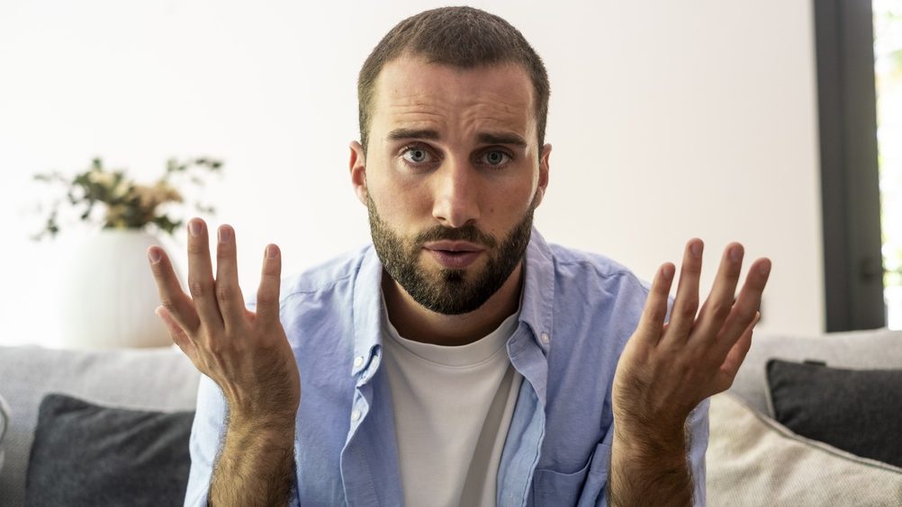 Bearded man sitting on a sofa at home raising his hands with a confused, frustrated expression as if asking a question or explaining something during a casual conversation or video call.