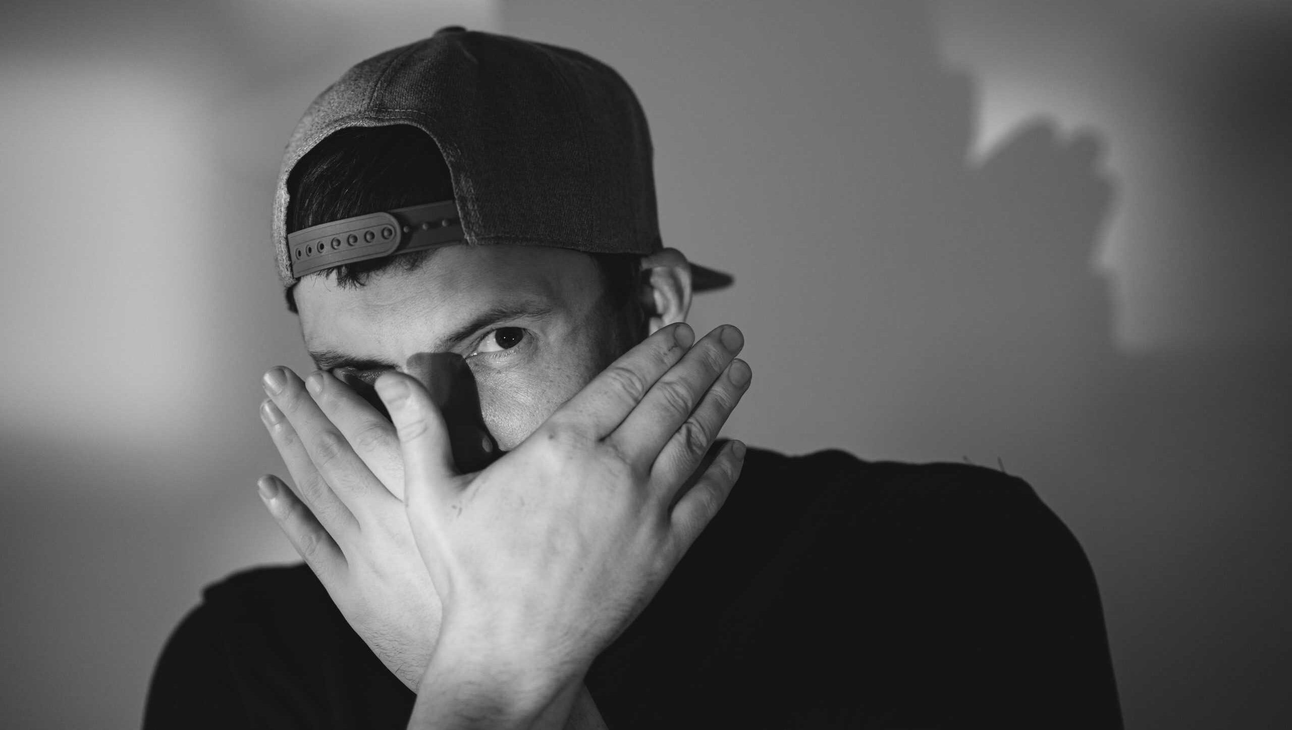 Young man wearing baseball cap covering face with hand. Candid portrait of male in shadowy lighting. Concept of hiding, mystery, shy expression and anonymous lifestyle in monochrome style.