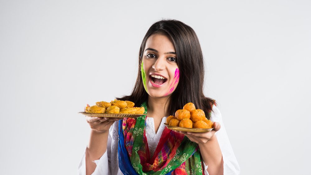 indian pretty young girl holding a plate full of sweet bundi laddu or gujiya and powder colour called gulal or gulaal on indian festival called Holi, isolated over white background, selective focus