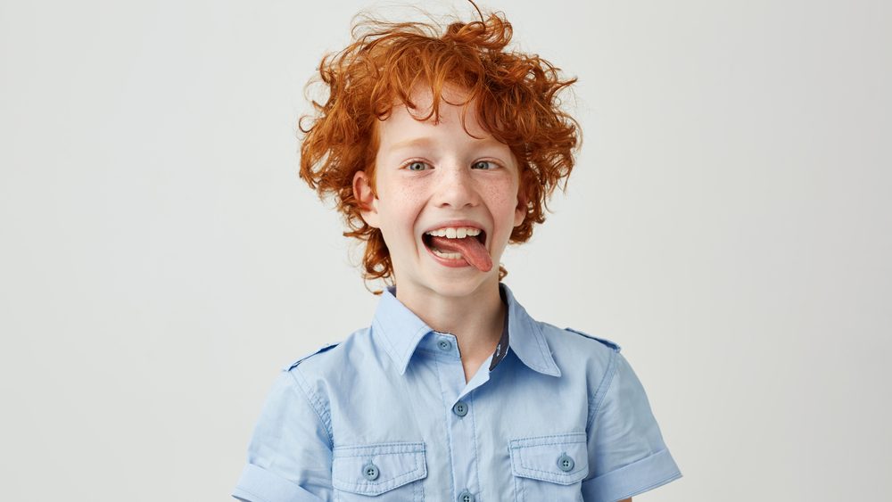 Portrait of silly little ginger boy in blue shirt with wild hair mowing eyes, smiling and showing tongue in camera,making funny faces.
