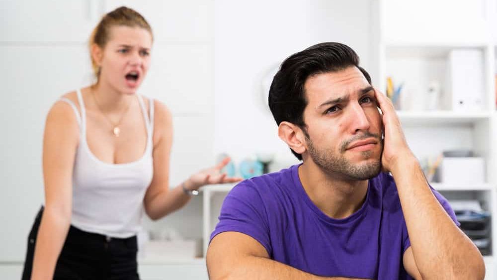 Young stressed man at home table with screaming wife behind