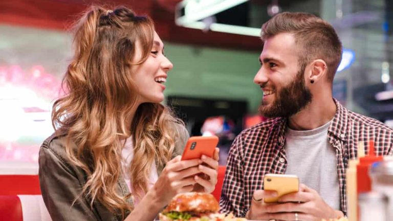 Photo of a cute pleased young loving couple in retro bright street food cafe using mobile phone.