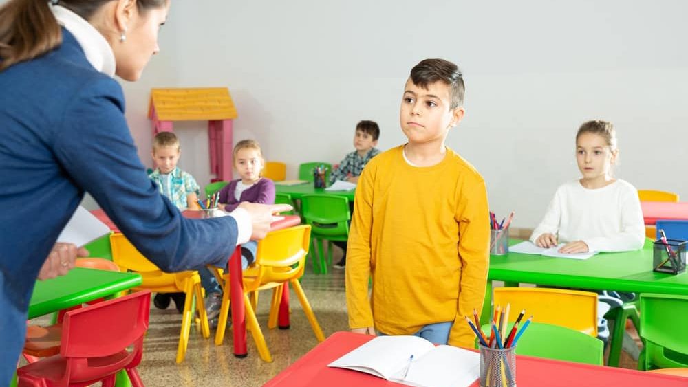 Upset boy listening reprimanding of strict female teacher during lesson in primary school