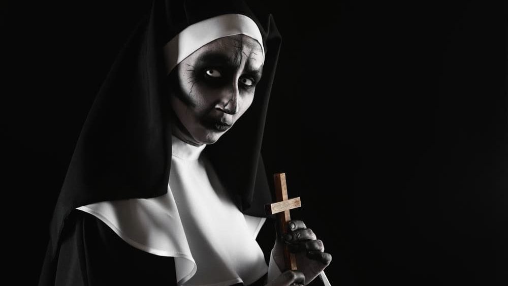Scary devilish nun with cross on black background, space for text. Halloween party look