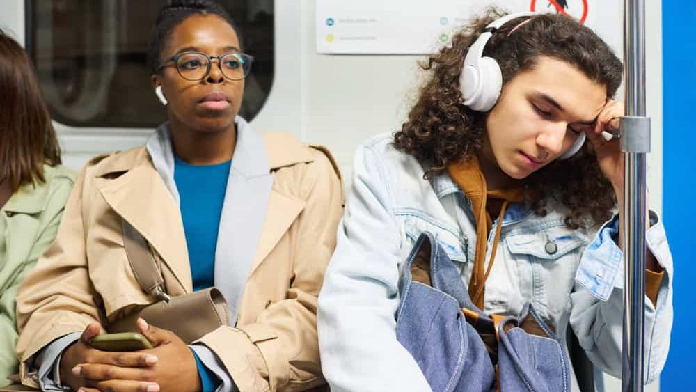 Young female passenger in earphones listening to music while sitting next to teenage guy in headphones napping against handrail in train