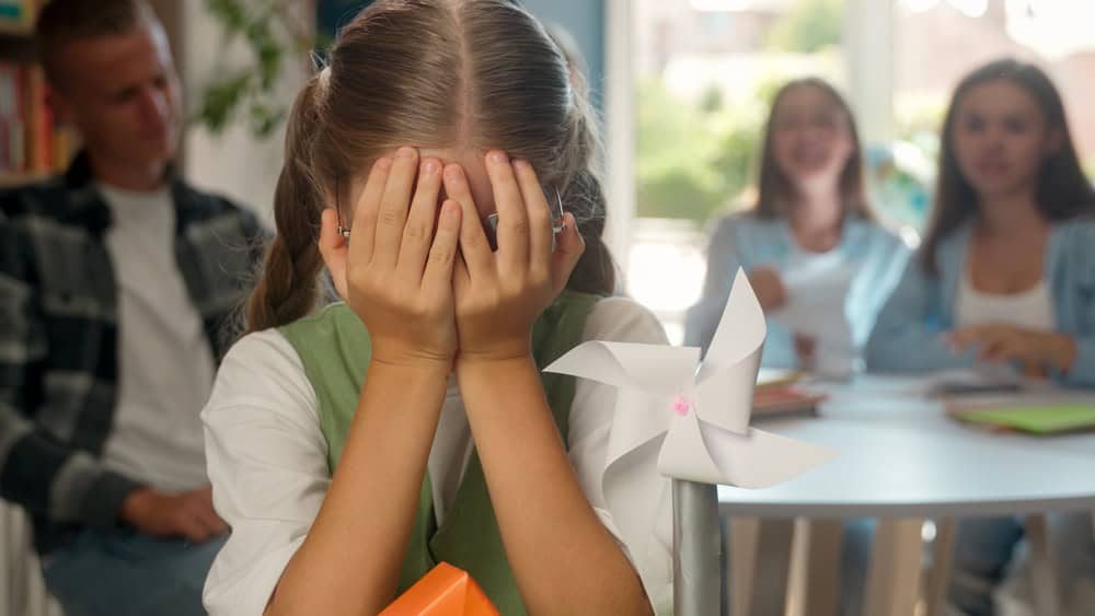 School bullying problem teenager violence victim Caucasian stressed crying girl child kid in class doing paper house craft art teenagers classmates throwing crumpled papers balls mock social trouble