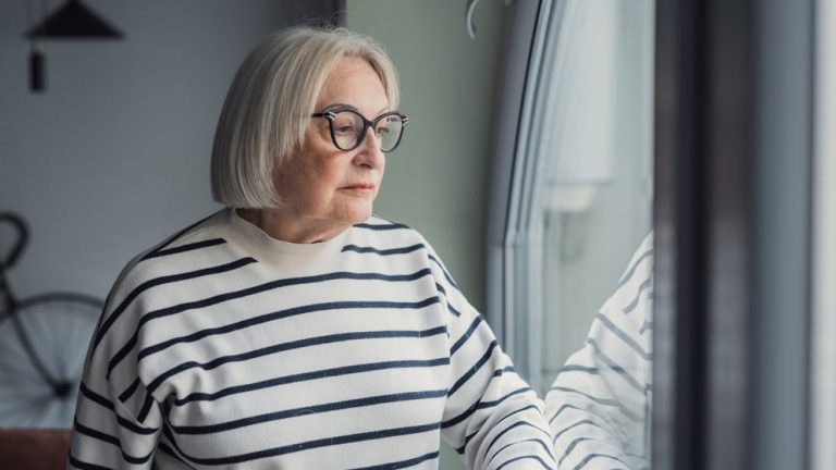 Elderly woman looks out the window with a worried expression. She reflects on her thoughts and feels concerned about her day while standing at home.