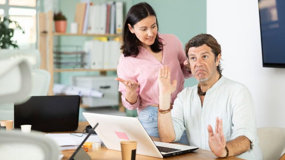 Adult woman sternly scolds adult male colleague who working on laptop in office