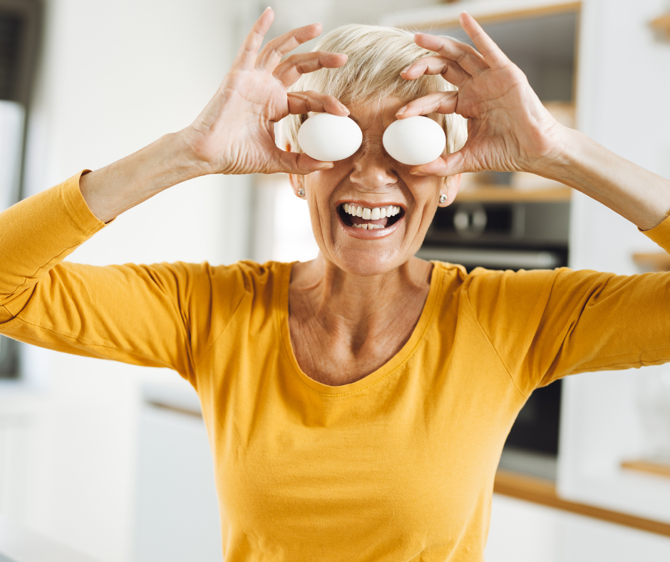 Mature woman in a yellow top holding two cooked eggs in her hand over her eyes standing in her white kitchen.