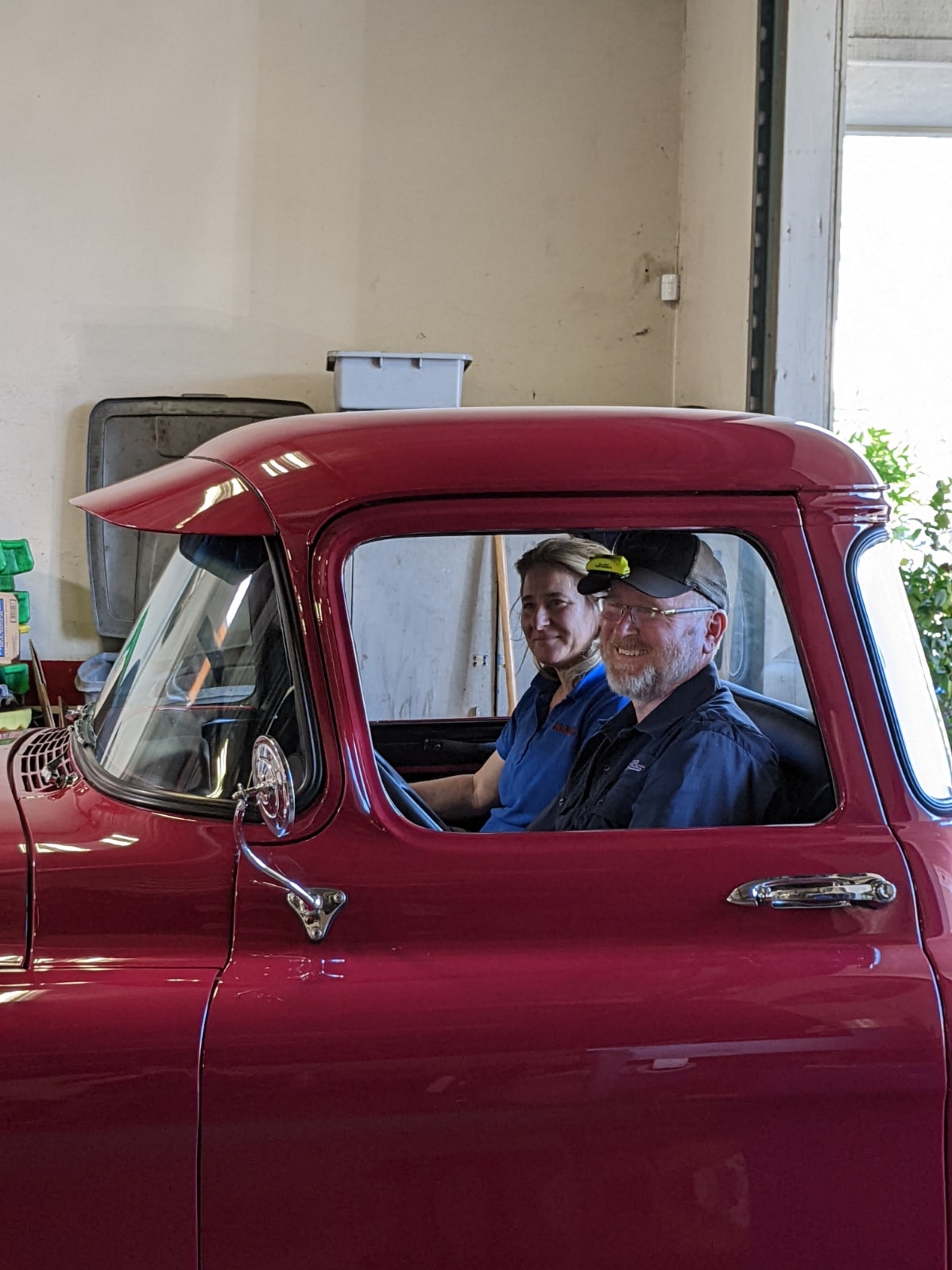 Vintage red truck with two smiling men inside, in a garage setting.
