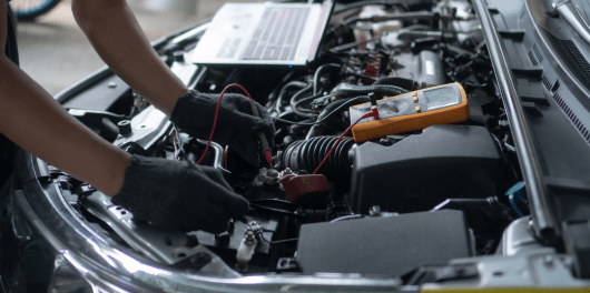 Auto repair technician testing car battery with multimeter in engine bay.