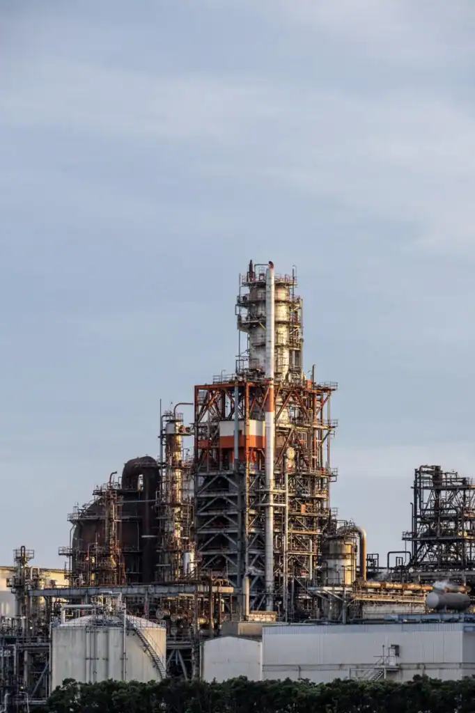 Refinery tower with complex piping and structures under a cloudy sky.