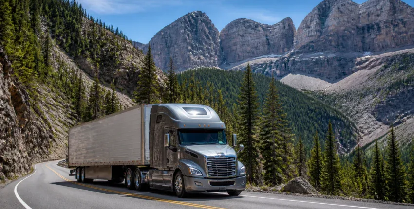 Semi truck driving through scenic mountain landscape with trees and rocky peaks.