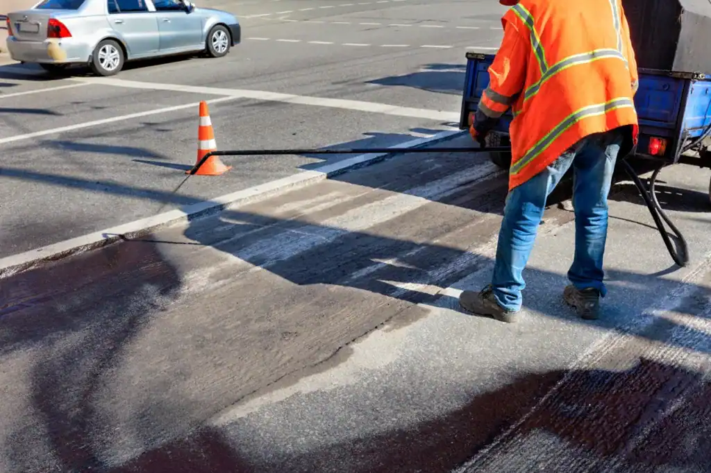 Worker in orange safety vest applying fresh asphalt to crosswalk pavement.