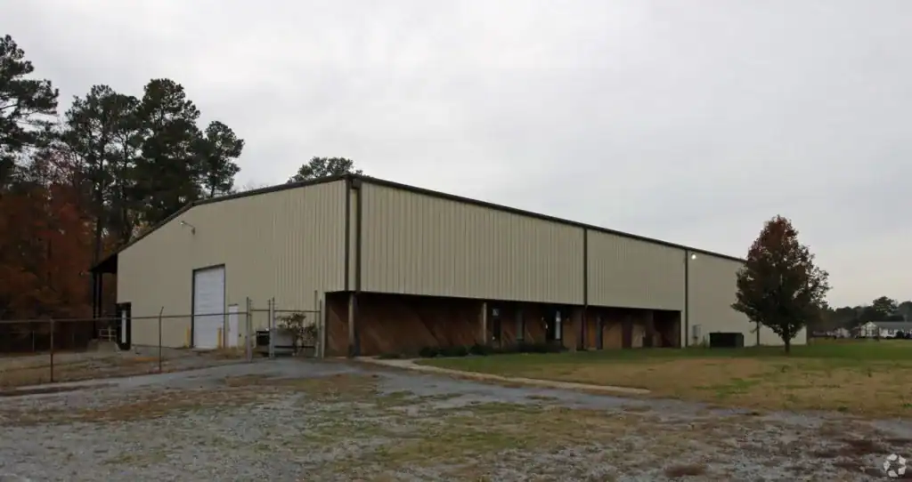 Large industrial warehouse with metal construction and wooden details, surrounded by open land and trees.
