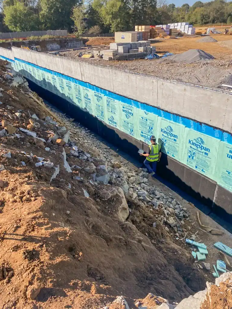 Worker inspecting foundation with insulation and building materials in background.