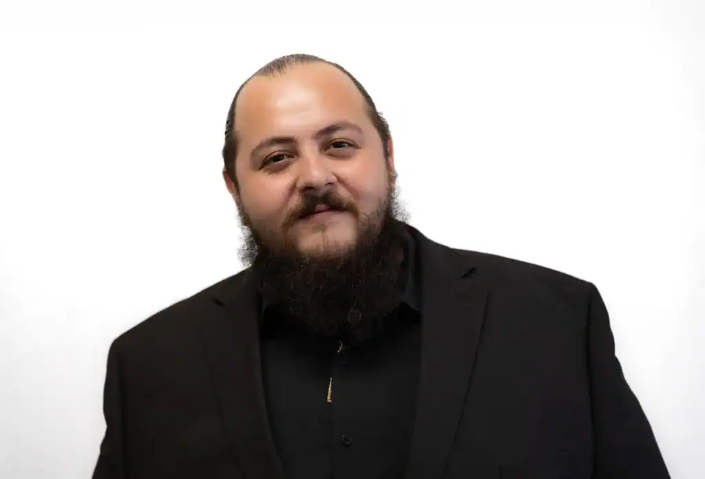 Portrait of a confident man with a beard wearing a black suit and shirt against a white background.