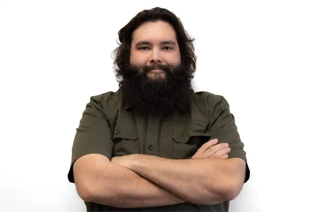 Male with long hair and beard, crossing arms confidently against a plain white backdrop.