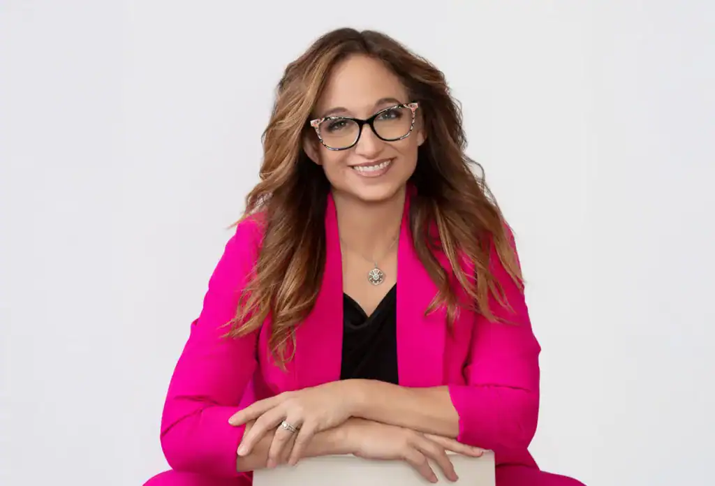 Stylish woman with glasses wearing a bright pink blazer, smiling confidently against a plain background.